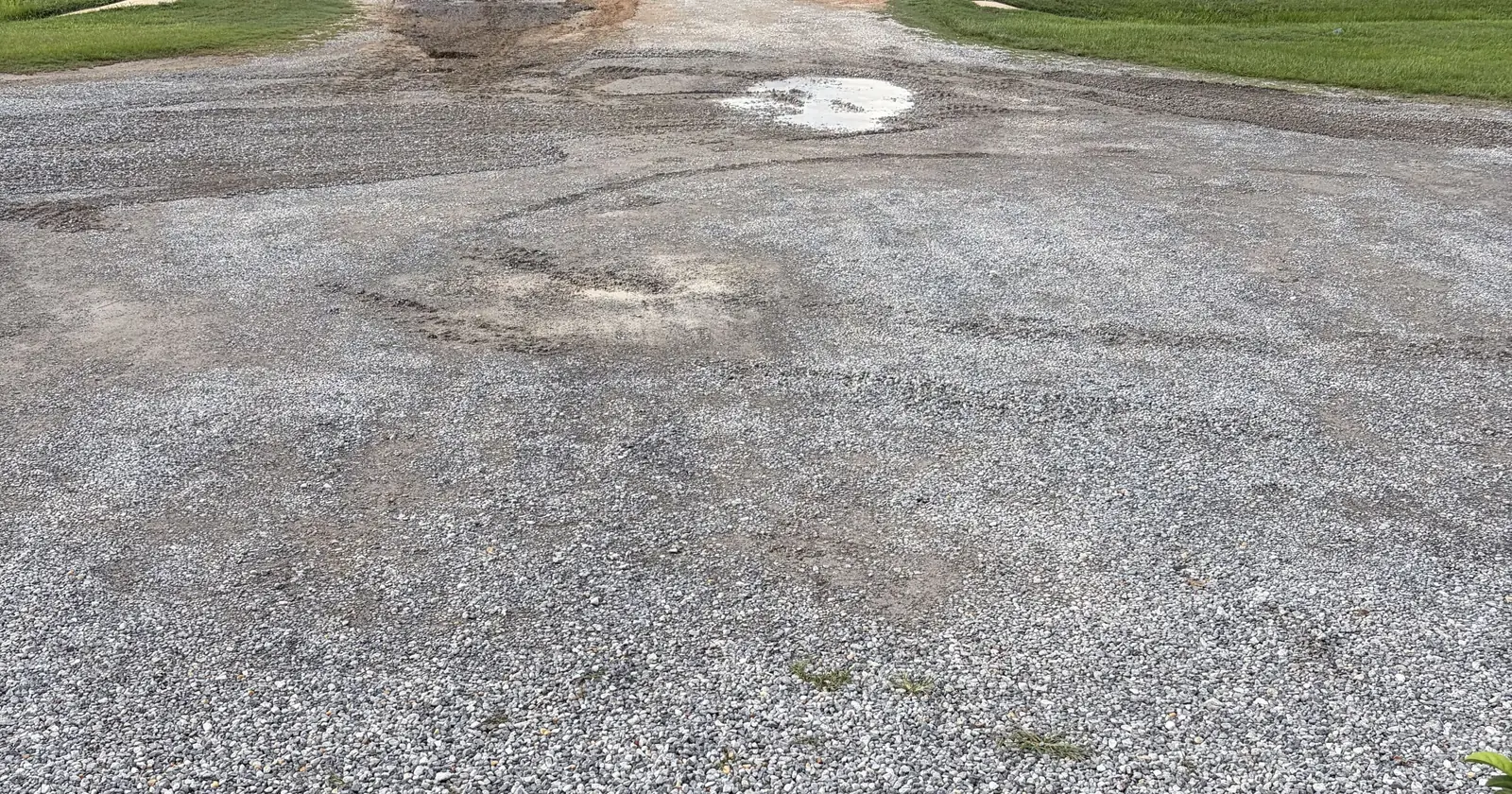 Gravel parking lot before regrading, with ruts, potholes, bare dirt and water standing near the road entrance