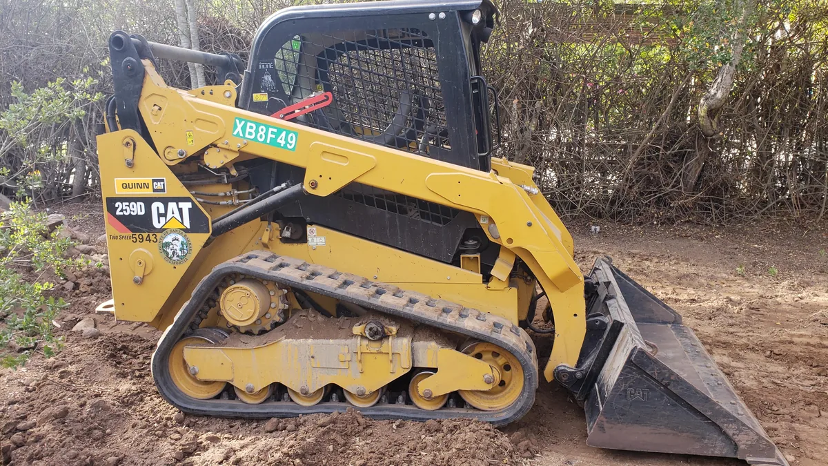 Compact track loader with a bucket on freshly graded dirt beside a line of brush